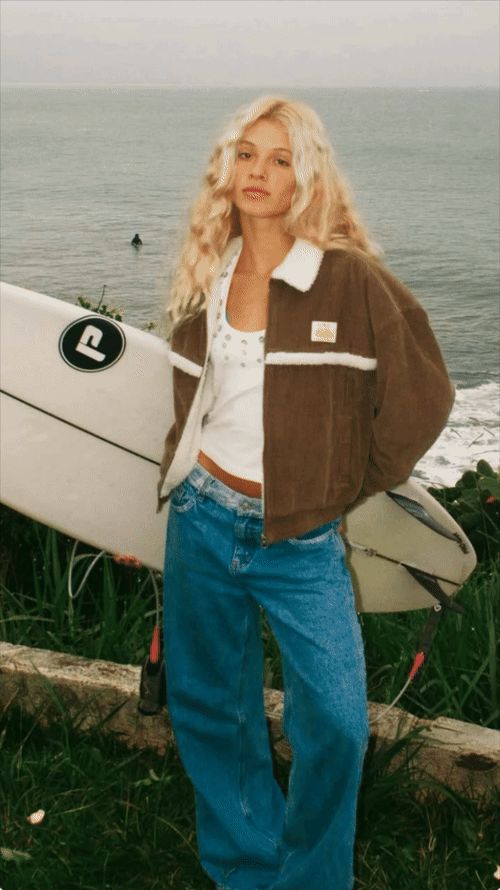 Animation of a model holding a surfboard by the ocean, with her outfit edited into different clothing-brand looks by Ideogram 4.5.
